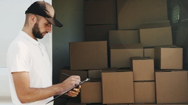 Deliveryman writing on clipboard next to stacked cardboard boxes in a van.